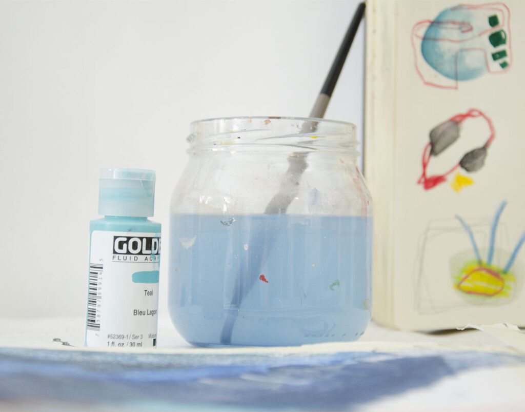 Studio image with acrylic paint and a a glass jag filled with blue water and a paint brush